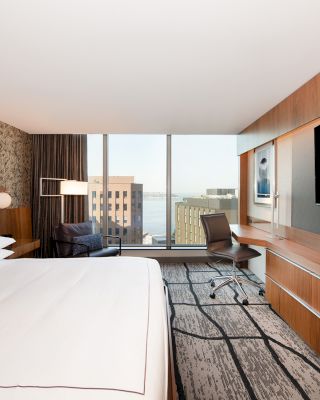 A modern hotel room with a large bed, TV, desk, and a city view through large windows. Contemporary design with a comfortable ambiance.