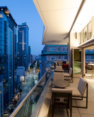 A modern balcony overlooks a city street lined with tall buildings, featuring outdoor furniture and tables, under an evening sky.