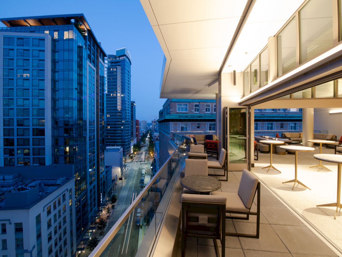 A modern outdoor balcony at dusk with tables, chairs, and city buildings in the background, featuring glass railings and urban views.