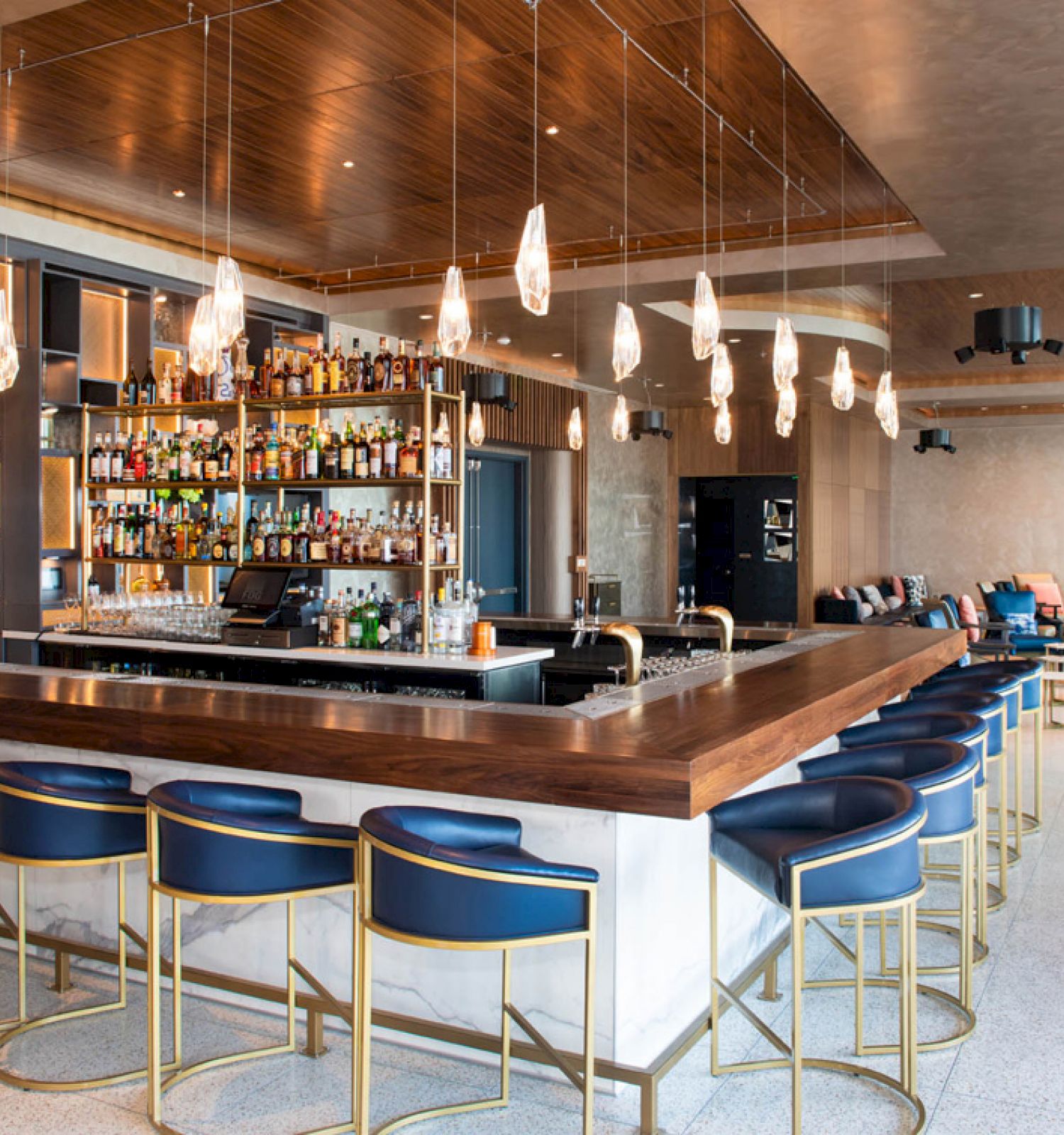 This image depicts a modern bar with a wooden counter, blue bar stools, shelves of liquor, pendant lights, and a comfortable seating area in the back.