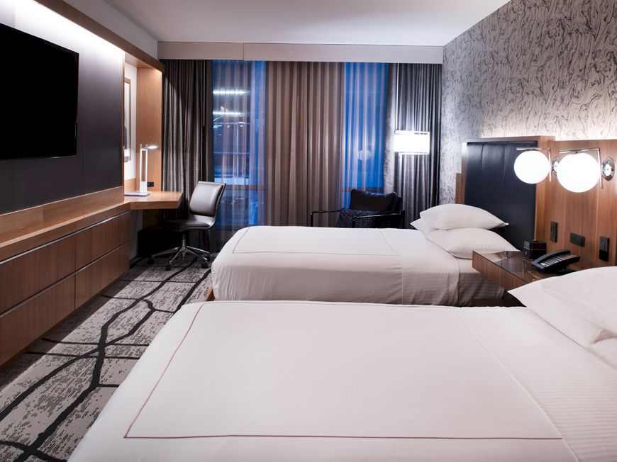 The image shows a modern hotel room with two neatly made beds, wall-mounted TV, desk, chair, and stylish decor with a patterned wall.