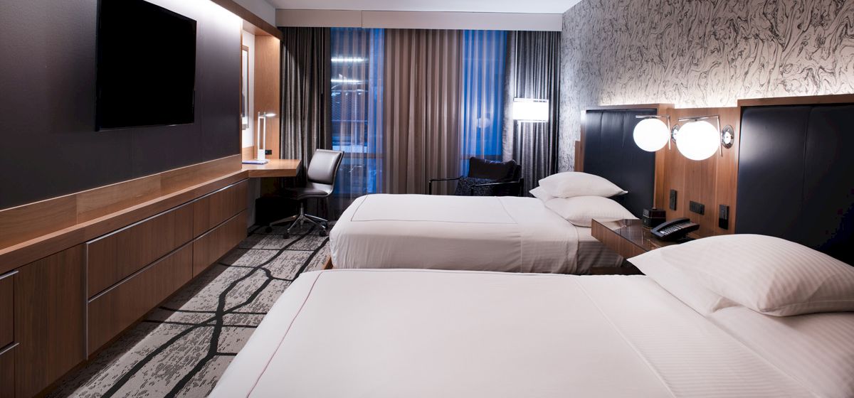 The image shows a modern hotel room with two neatly made beds, wall-mounted TV, desk, chair, and stylish decor with a patterned wall.