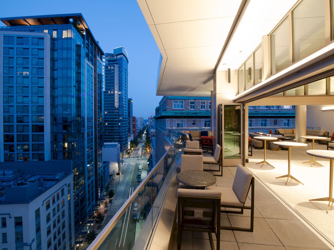 A modern balcony with seating and tables, overlooking a city street lined with tall buildings at dusk, featuring large glass windows and urban views.