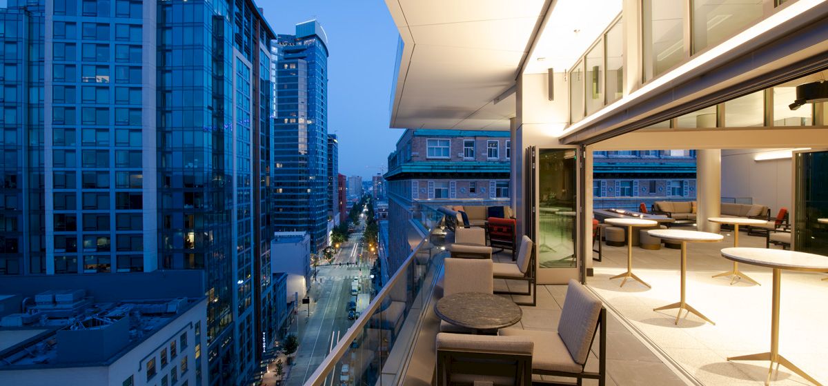 A modern balcony with seating and tables, overlooking a city street lined with tall buildings at dusk, featuring large glass windows and urban views.