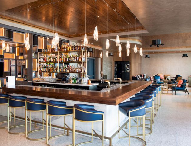 The image depicts a modern bar with a wooden counter, multiple hanging lights, blue cushioned bar stools, and a well-stocked shelf of bottles.