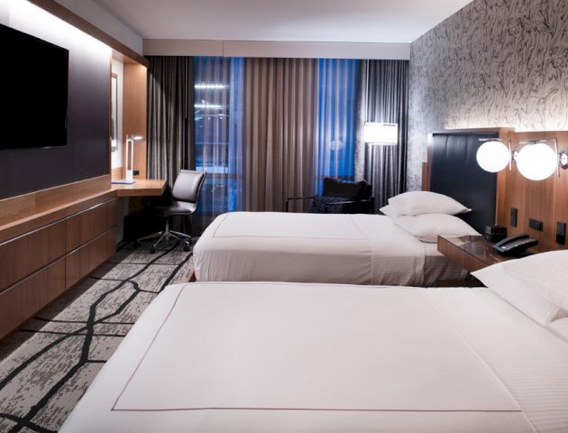 A modern hotel room with two neatly made beds, a large TV, a desk and chair, and ambient lighting with patterned wallpaper creates a cozy ambiance.