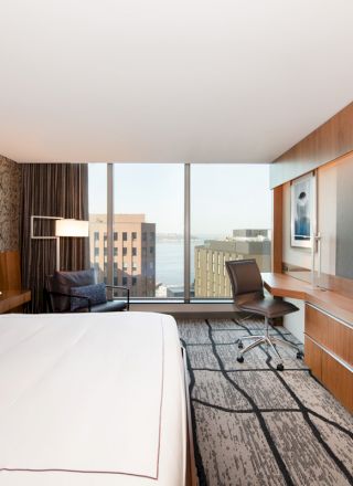 A modern hotel room featuring a large bed, a TV, a desk with a chair, and a city view through a large window, is shown.