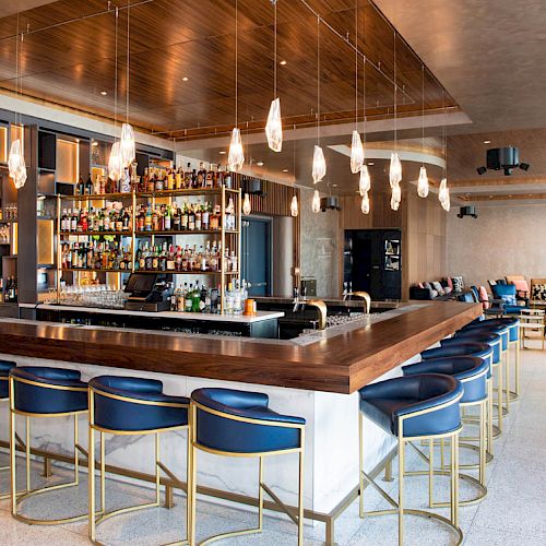 This image shows a modern bar with a wooden counter, stylish barstools, a variety of bottles on shelves, and hanging lights in a spacious setting.