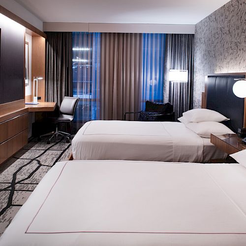 The image shows a modern hotel room with two double beds, a desk with a chair, a flat-screen TV on the wall, and large curtained windows.