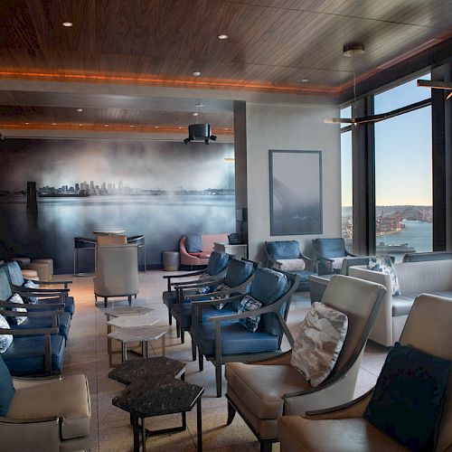 A modern lounge area with ample seating, a mural on the wall, and large windows offering scenic views of a waterfront cityscape at sunset.