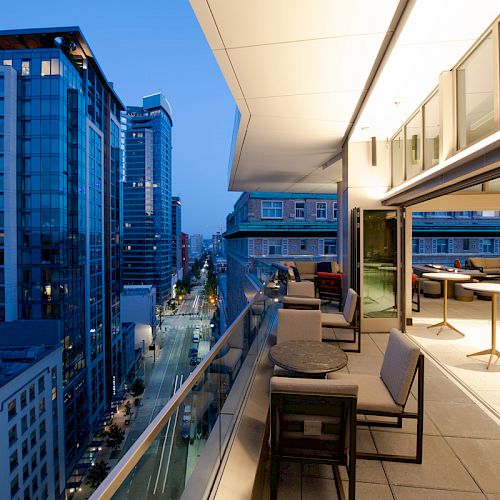 A modern balcony with seating and tables overlooks a city street lined with tall buildings at dusk. The view captures an urban evening ambiance.