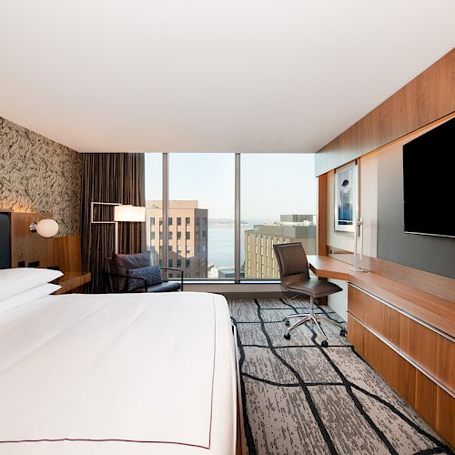 A modern hotel room with a large bed, a wall-mounted TV, a desk, and a chair. There is a view of buildings and water through the window.
