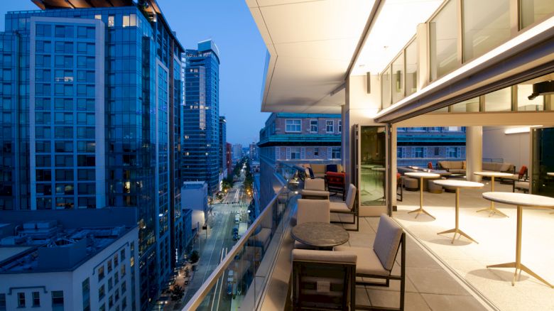 A modern balcony overlooks a bustling city skyline at dusk, featuring comfortable seating and tables, illuminated by indoor and outdoor lighting.