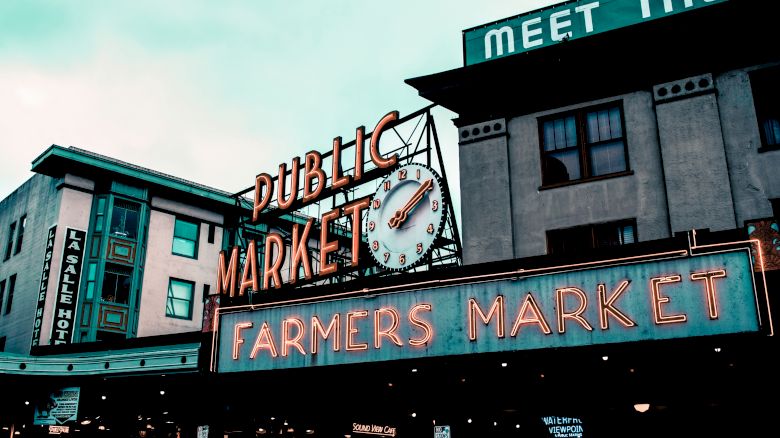 A neon sign reads "Public Market" and "Farmers Market" below a clock. Nearby buildings have signs and windows. The sky is cloudy.