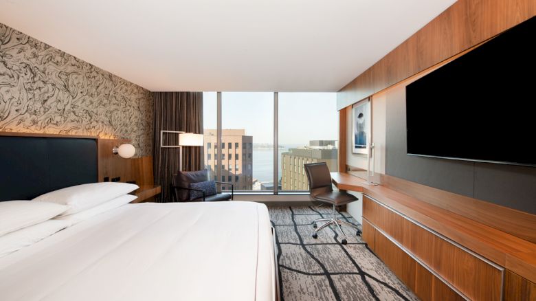 The image features a modern hotel room with a large bed, a desk, a chair, a wall-mounted TV, and a view of cityscape through wide windows.