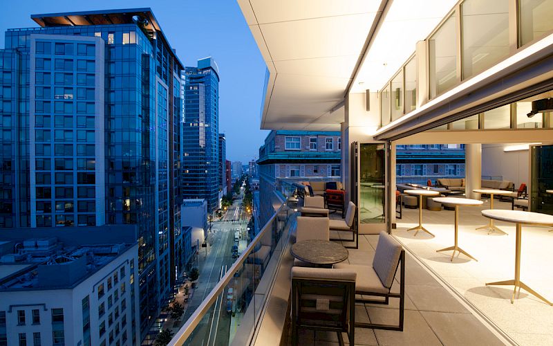 A modern balcony with tables and chairs overlooks a cityscape of tall buildings at dusk, featuring large windows and a serene ambiance.
