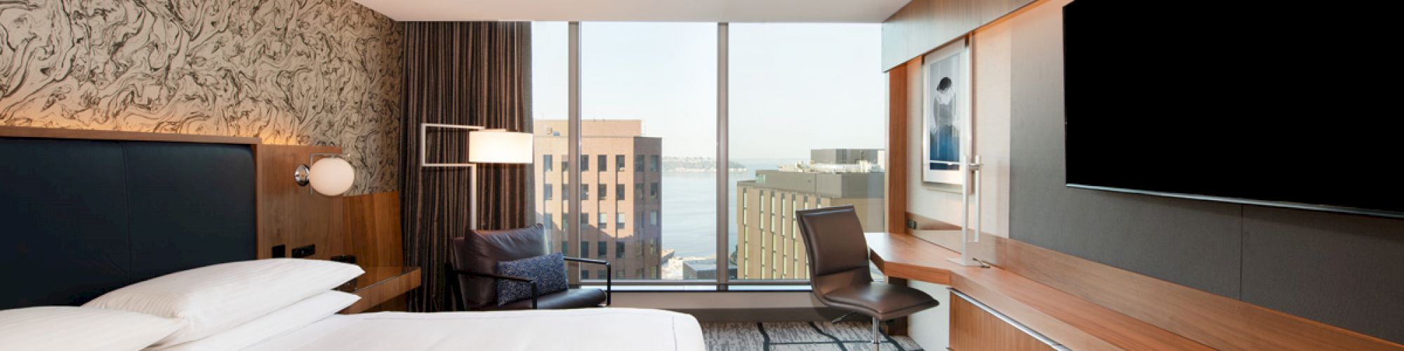 A modern hotel room featuring a large bed, a desk with a chair, a wall-mounted TV, and a city view through ceiling-to-floor windows.