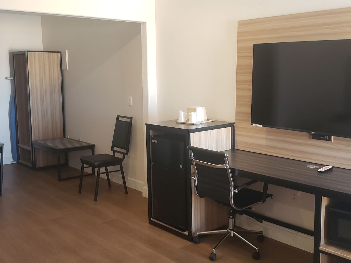 A tidy hotel room office setup with a desk, chair, TV, drawer cabinet, and a doorway with a small box behind it.
