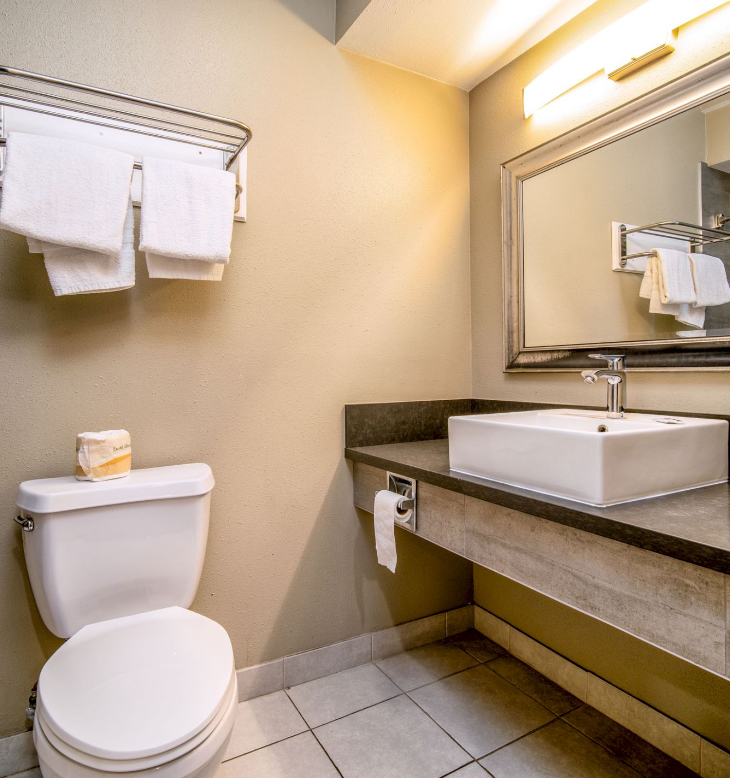 A clean bathroom with a toilet, sink, large mirror, towel rack, and tiled floor. Towels are neatly folded on the rack.