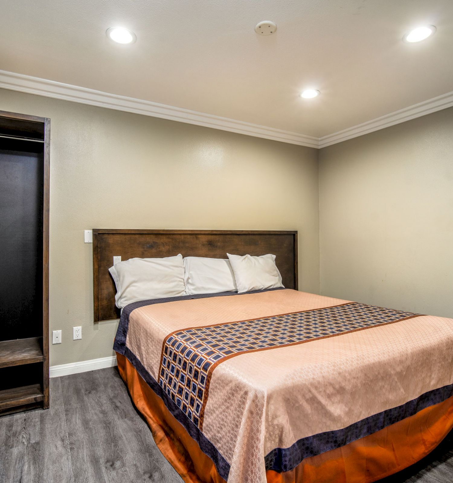 A minimalist bedroom with a large bed, simple bedding, wood flooring, an open wardrobe, and recessed lighting on the ceiling.