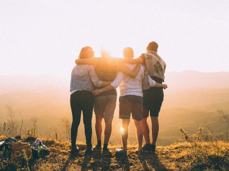 A group of four people stands on a hilltop, arms around each other, with a beautiful sunlit valley stretching out beneath them at sunset.