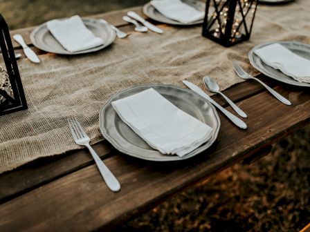 A rustic table setting with plates, cutlery, and white napkins on a burlap runner, with lanterns as centerpieces, ready for an outdoor meal.