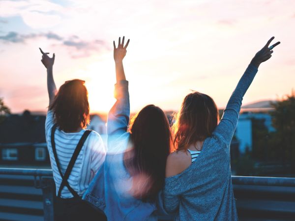 Three individuals stand with their backs to the camera, raising their arms and making peace signs as the sun sets, creating a serene atmosphere.