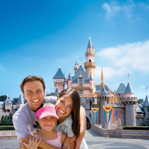 A smiling family poses in front of a fairy-tale castle at an amusement park, with clear blue skies in the background.