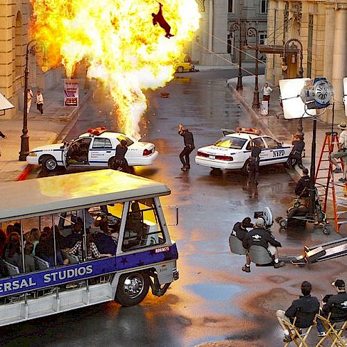 A film set with an explosion, NYPD cars, production crew, and a Universal Studios bus full of people.